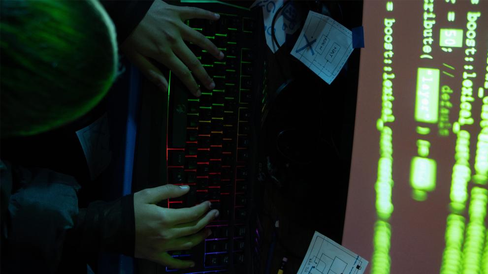 A photograph of a child’s hands as if typing on a colourful keyboard. On the left side we see a part of the child’s head photographed from the top and on the right we see a computer screen showing a green coloured code. 