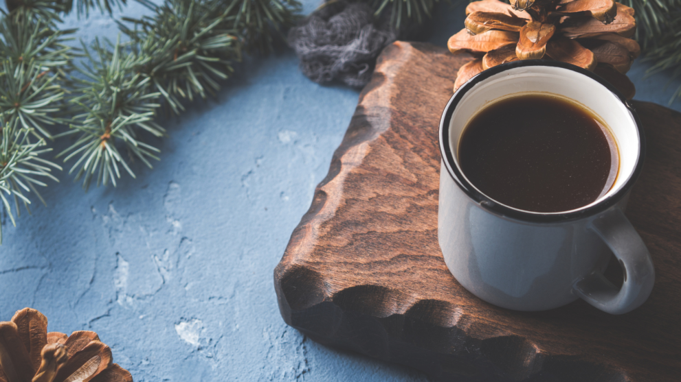 A photograph of coffee in a cup on a wooden board with pine tree branches.