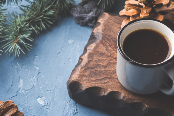 A photograph of coffee in a cup on a wooden board with pine tree branches.